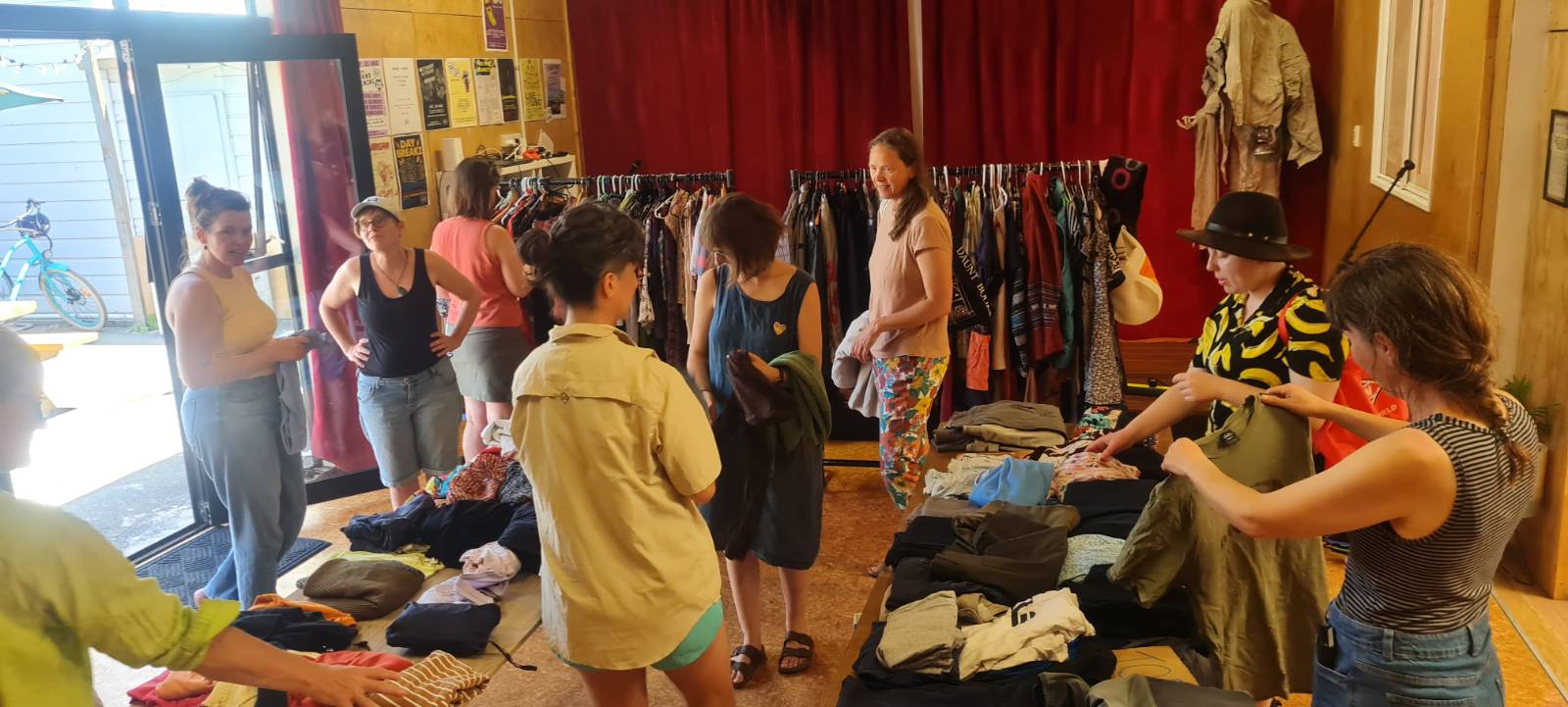 A group of Timebank Whakatū members and friends looking over clothes at the clothes swap. There are approximately ten people going through clothing spread across two tables and a rack in the background.